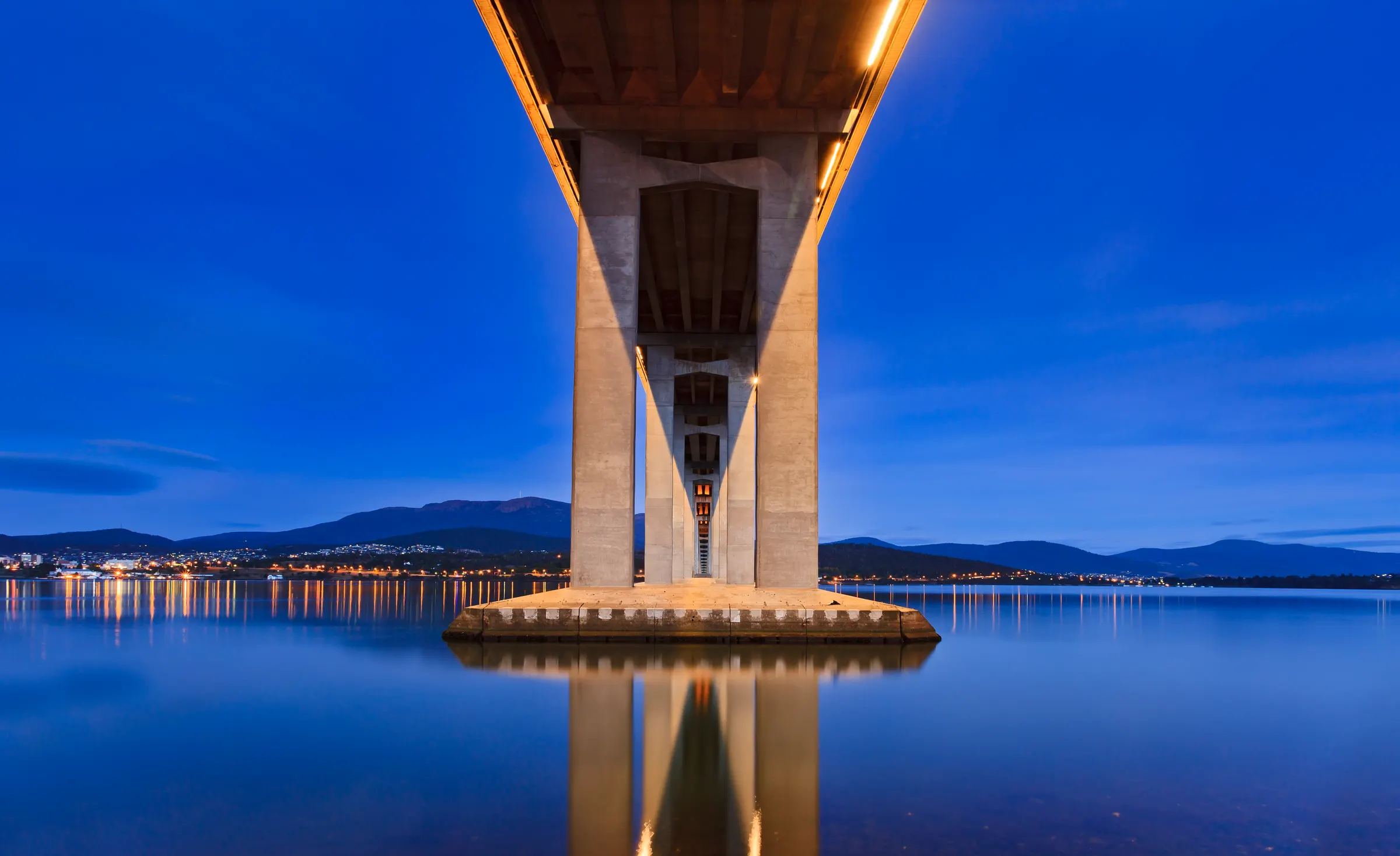 The Tasman Bridge seen from directly below at blue hour, a symmetric concrete pylon rising out of still water beneath a deep blue sky, with Hobart's lights on the horizon